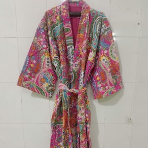 Könnte beinhalten: Langer, rosa Paisley-Muster-Bademantel mit passendem Gürtel. Der Bademantel hat ein farbenfrohes Blumenmuster mit Rosa-, Blau-, Grün- und Gelbtönen. Der Bademantel hängt an einer weiß gefliesten Wand.