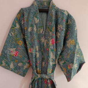 May include: A teal-colored, floral-patterned robe with a matching belt. The robe features a V-neck and wide sleeves. The floral design includes pink, yellow, and white accents, with white stitching throughout. The robe is made of a soft, lightweight fabric.