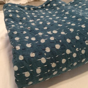 May include: A folded teal-colored textile with a repeating pattern of white polka dots. The fabric is stitched with white and dark blue thread, creating a textured appearance. The textile is likely a blanket or quilt.