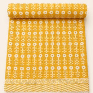 Könnte beinhalten: Aufgerollte gelbe Steppdecke mit weißem Blumenmuster. Die Steppdecke hat ein sich wiederholendes Design aus stilisierten Blumen und Stielen. Ein dekorativer Rand mit Blumen- und Streifenmuster ist am Rand sichtbar.