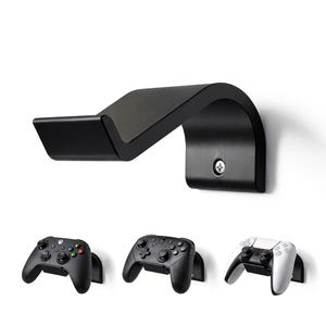 Könnte beinhalten: Ein schwarzer, an der Wand montierter Gaming-Controller-Halter mit geschwungenem Design. Darunter werden drei Gamecontroller angezeigt: ein schwarzer Xbox-Controller, ein schwarzer Controller und ein weiß-schwarzer PlayStation-Controller.