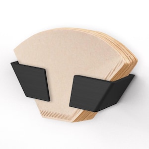 May include: A stack of beige coffee filters held in a black, angular holder. The holder has two angled arms that cradle the filters, creating a modern and minimalist design. The filters are made of paper and have a textured appearance.