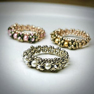 May include: Three handcrafted beaded rings arranged in a cluster. The front ring features metallic gold beads and white pearls. The back left ring combines metallic gold beads with pink pearls and green accents. The back right ring features gold beads.