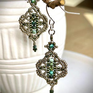 May include: A pair of dangle earrings featuring a diamond-shaped woven bead design. The earrings are crafted with metallic bronze, olive green, and turquoise beads, with a central vertical row of turquoise stones and silver-toned metal hooks.