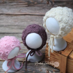 Könnte beinhalten: Drei gestrickte Babyhauben in Rosa, Lila und Creme, jeweils auf einem weißen Schaufensterkopf präsentiert. Die Hauben haben ein strukturiertes Muster und sind mit passenden Bändern gebunden. Der Hintergrund ist eine Holzoberfläche.