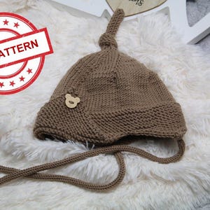 Puede incluir: Un gorro de punto marrón con un botón en forma de oso y un cordón largo anudado. El gorro está sobre una superficie blanca y esponjosa.