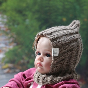 Könnte beinhalten: Eine braune gestrickte Baby-Mütze mit Ohrenklappen und einem kleinen weißen Etikett mit der Aufschrift "hand made". Die Mütze wird von einem Baby mit blauen Augen und einem rosa Shirt getragen.
