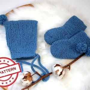 May include: A blue knitted baby hat and booties set with pom poms. The hat has a tie at the back. The set is on a white fluffy surface.