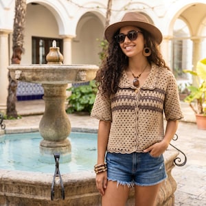 Puede incluir: Una camisa de crochet de manga corta en color beige y marrón con cuello y dobladillo festoneados, combinada con pantalones cortos de mezclilla azul, un sombrero fedora marrón, gafas de sol oscuras, un collar de cuentas con colgante y pulseras.