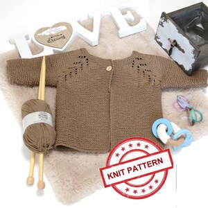 May include: A brown knitted baby cardigan with a button closure and lace detailing on the shoulders. The cardigan is laid out on a white faux fur surface with wooden knitting needles and yarn.