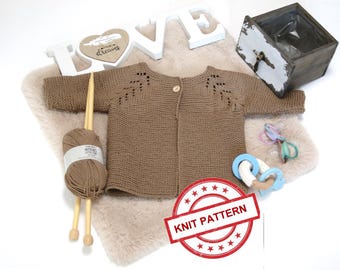 May include: A brown knitted baby cardigan with a button fastening and lace detailing on the shoulders. The cardigan is laid out on a white faux fur surface with wooden knitting needles and yarn.