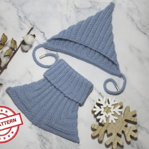 Könnte beinhalten: Hellblaues Strickmützen- und Halstuch-Set. Die Mütze ist spitz mit Ohrenklappen und Bindebändern. Der Halswärmer ist ein geripptes Rohr. Ein Holzschneeflocke und ein Baumwollzweig sind ebenfalls im Bild. Der Text "KNIT PATTERN" befindet sich in einem roten Kreis.