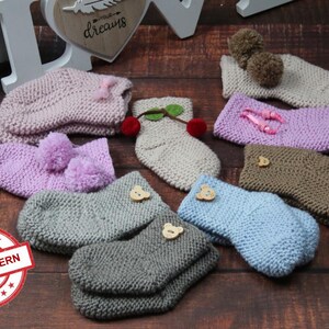 May include: A collection of knitted baby booties in various colors, including pink, purple, beige, brown, and blue. Some booties have pom-poms, while others have decorative buttons. The booties are arranged on a wooden surface with a sign that reads "Knit Pattern".