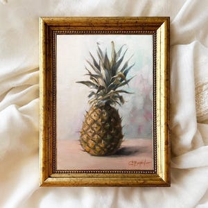 May include: A framed painting of a pineapple with a brown and green color scheme. The pineapple is sitting on a light pink surface. The painting is in a gold frame with a beaded border.