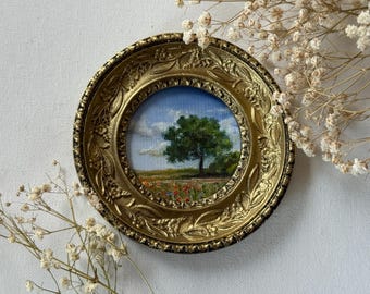 May include: A miniature landscape painting in a round, ornate gold frame. The artwork depicts a tree in a field with a blue sky and clouds. The frame has detailed floral and leaf patterns. Dried flowers are arranged around the frame.