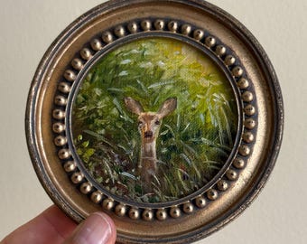 May include: A circular painting of a deer head peeking through tall green grass, enclosed in a round, antique-style bronze frame with a beaded edge. The artwork features shades of brown, tan, and vibrant green, capturing the animal in a natural setting.