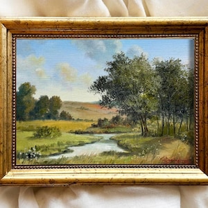 May include: Oil painting of a landscape scene with a stream, trees, and a field. The artwork is framed in a gold-colored, ornate frame. The sky is blue with some clouds.