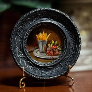 May include: A miniature painting of a meal, featuring a steak, fries, and salad, presented on a small plate. The painting is set within a decorative, black, circular frame, supported by a gold-colored stand. The frame has an ornate floral design.
