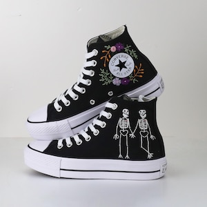 May include: Black high-top sneakers with white laces and soles. One shoe features floral embroidery and the Converse All Star logo. The other shoe has a skeleton design. The shoes are stacked on top of each other.