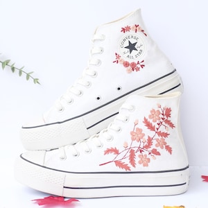 May include: White high-top sneakers with floral embroidery. The sneakers feature a black stripe along the sole and a classic Converse All Star logo. The floral embroidery includes pink and red flowers and leaves.