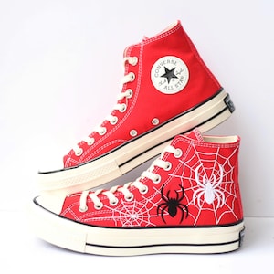 Puede incluir: Zapatillas altas rojas con cordones blancos y suela negra. Una zapatilla presenta un diseño de telaraña blanca con una araña negra y una araña blanca. La otra tiene el logo de Converse All Star.