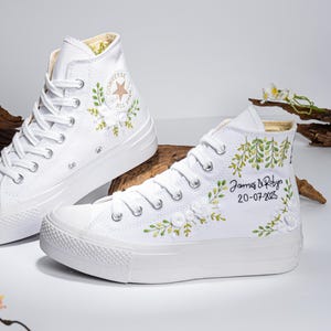 Può includere: Sneakers Converse bianche alte con ricamo floreale verde e bianco. La scarpa destra ha il testo "James & Robyn 20-07-2025" ricamato.