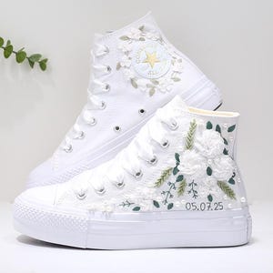 May include: White high-top sneakers with floral embroidery. The sneakers feature white laces, a rubber sole, and decorative embellishments including white flowers, green leaves, and small pearls. The date "05.07.25" is embroidered on the side.
