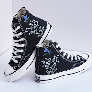 Könnte beinhalten: Schwarze High-Top-Canvas-Sneaker mit weißen Sohlen und gestickten Blumendesigns, darunter Libellen, auf beiden Schuhen.
