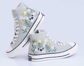 Puede incluir: Un par de zapatillas de lona de caña alta de color gris claro con margaritas blancas bordadas, tallos verdes y trigo amarillo. Las zapatillas tienen un diseño de perro pug bordado en negro.