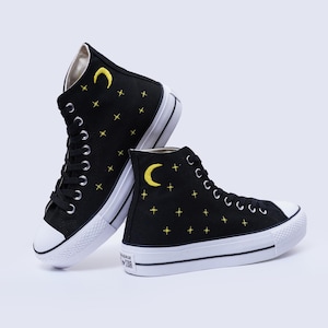 May include: Black high-top sneakers with white soles and laces. The sneakers feature yellow embroidered stars and crescent moons. The brand name is visible on the white sole.