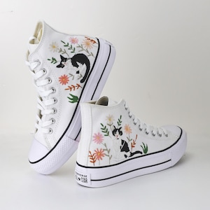May include: White high-top canvas sneakers with black and white embroidered cats and floral designs. The shoes have a black trim and a white rubber sole. Converse All-Star is printed on the side.
