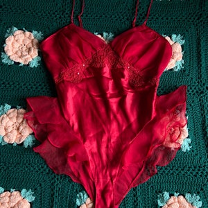May include: A red satin bodysuit with a lace trim and a ruffled hem. The bodysuit is on a green crocheted background.