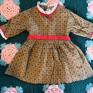 Puede incluir: Un vestido vintage de estampado amarillo, rojo y verde con una cintura de cinta roja y ribete de encaje blanco alrededor del cuello y las mangas.