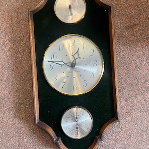 May include: A wooden framed wall clock with a gold trim and a green velvet background. The clock face has Roman numerals and a gold-colored hour and minute hand. The clock also has two smaller dials, one for the barometer and one for the thermometer.