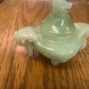 May include: A pale green jade teapot with a dragon-like spout and handle. The teapot has a rounded body and sits on three small feet. The lid is topped with a dragon head. The teapot is likely a decorative item.
