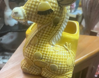 May include: A yellow ceramic giraffe planter with brown spots and a checkered pattern. The planter has a square opening on the back for planting.
