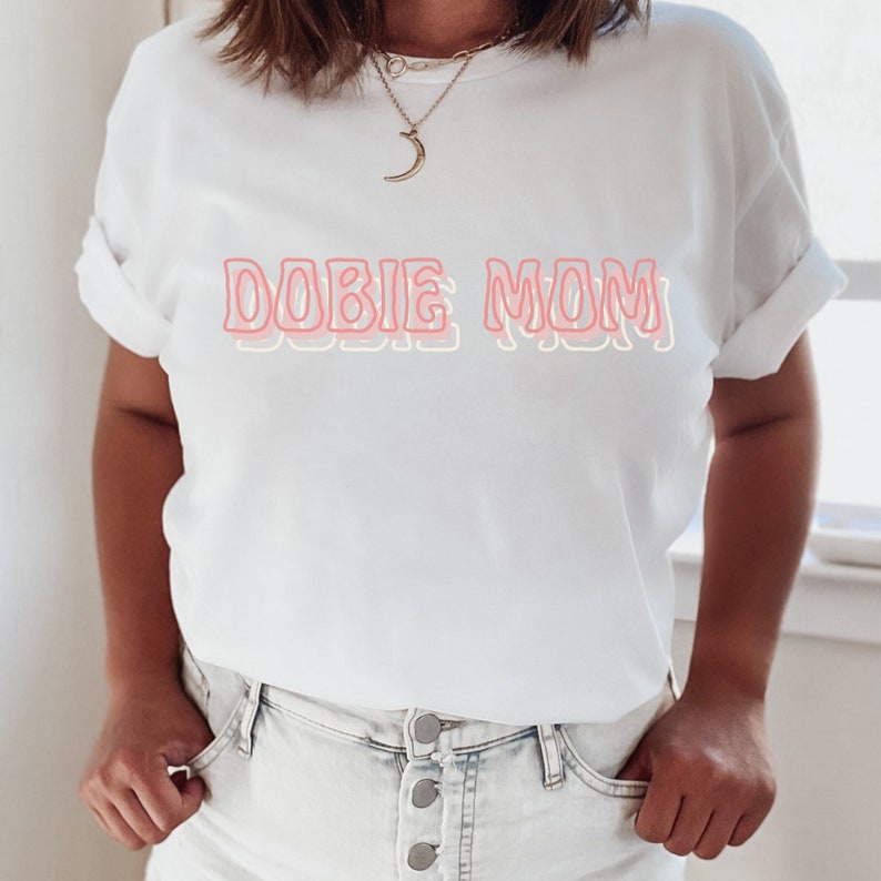 May include: White t-shirt with the text "DOBIE MOM" in pink and yellow outlined letters.