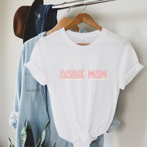 May include: White t-shirt with the text "DOBIE MOM" in pink outlined letters.