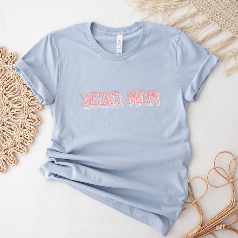 May include: Light blue t-shirt with the text "DOBIE MOM" in pink outlined letters.