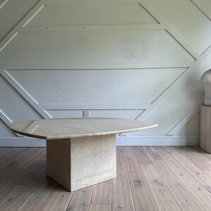 May include: A light beige, triangular coffee table with a solid, rectangular base. The table is made of a light-colored stone. A white sculpture of a seated figure is on a matching pedestal. The room has light wood floors.