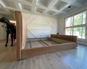 May include: A modern bed frame with a light brown leather headboard and footboard. The headboard has vertical stitched detailing. The bed frame is set on a light wood floor in a room with large windows and a decorative ceiling.
