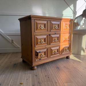 May include: A wooden dresser with six drawers, each with a square panel and gold-colored hardware. The dresser is made of light brown wood and sits on short, rounded legs. The room has a hardwood floor.