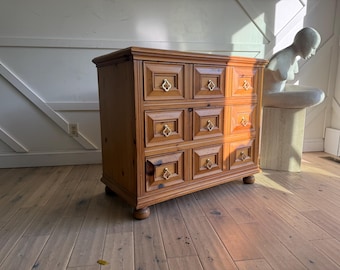 May include: A wooden dresser with six drawers, each with a square panel and gold-colored hardware. The dresser is made of light brown wood and sits on short, rounded legs. The room has a hardwood floor.
