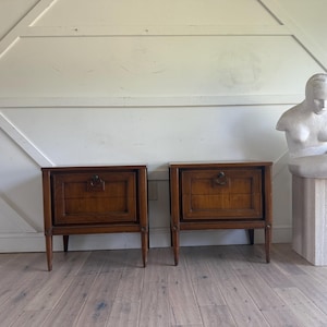 May include: Two matching wooden nightstands with a warm brown finish. Each nightstand features a drawer with a decorative handle and slender legs. A white sculpture of a seated figure is on a stone pedestal.