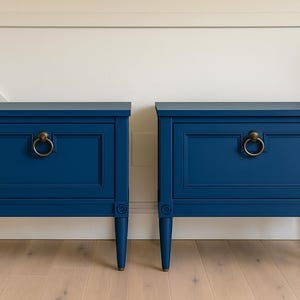 May include: Two matching navy blue nightstands with brass ring pulls. Each nightstand has a rectangular design with a recessed panel on the drawer front and sits on four tapered legs. The background is a white wall and light wood floor.