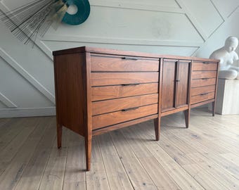 May include: A long, dark wood dresser with six drawers and two cabinet doors. The dresser has a simple, mid-century modern design with sleek lines and metal hardware.
