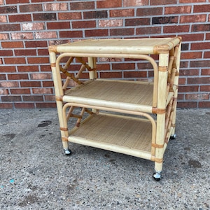 May include: A light-colored, three-tiered bamboo rolling cart with woven shelves. The cart has a rectangular shape with rounded corners and is supported by four small wheels. The frame is made of bamboo and has a natural finish.
