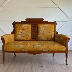May include: A vintage loveseat with a wooden frame and ornate details. The seat and back cushions are covered in a mustard yellow fabric with a pattern of white and gray cranes. The loveseat has curved arms and turned legs.