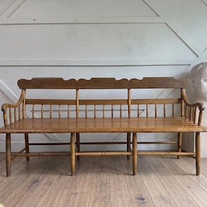 May include: A long wooden bench featuring a high backrest with a scalloped top edge, vertical spindle supports, and curved armrests. The bench is constructed from light brown wood with a smooth finish, supported by six legs connected by stretchers.