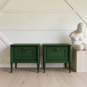 May include: Two dark green nightstands with brass ring pulls. Each nightstand has a single drawer and decorative legs. A white sculpture sits on a wooden block to the right of the nightstands. The background is a white wall.